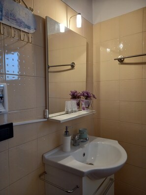 A bathroom with a white sink, a mirror, a towel rack, and a shelf with toiletries. - Adam Venice 2 (Venezia)