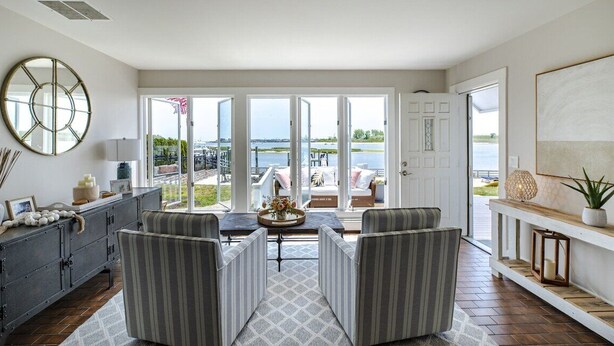 A spacious living room with a large window overlooking a body of water, a dining area with striped chairs, and a decorative console table. - Riverfront Shore House w/ Ocean View, Private Beach, 6 Bedrooms and Lots of Toys (Brielle)
