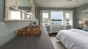 A bedroom with a large window, a ceiling fan, and a wicker basket on a wooden table. - Riverfront Shore House w/ Ocean View, Private Beach, 6 Bedrooms and Lots of Toys (Brielle)