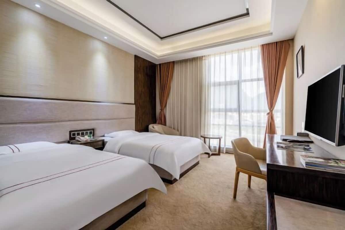A hotel room with two beds, a desk, a chair, a TV, and a large window with curtains.
