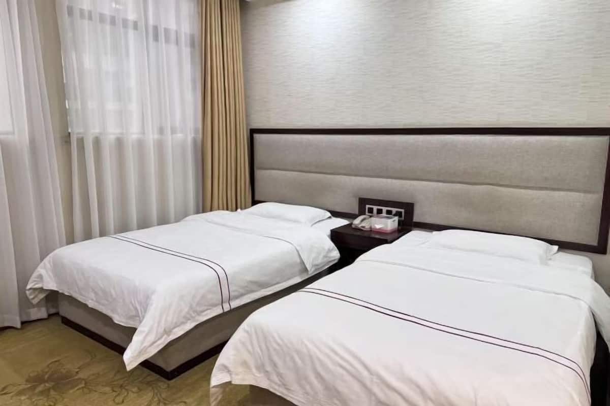 A hotel room with two beds, a nightstand, and a window with sheer curtains.