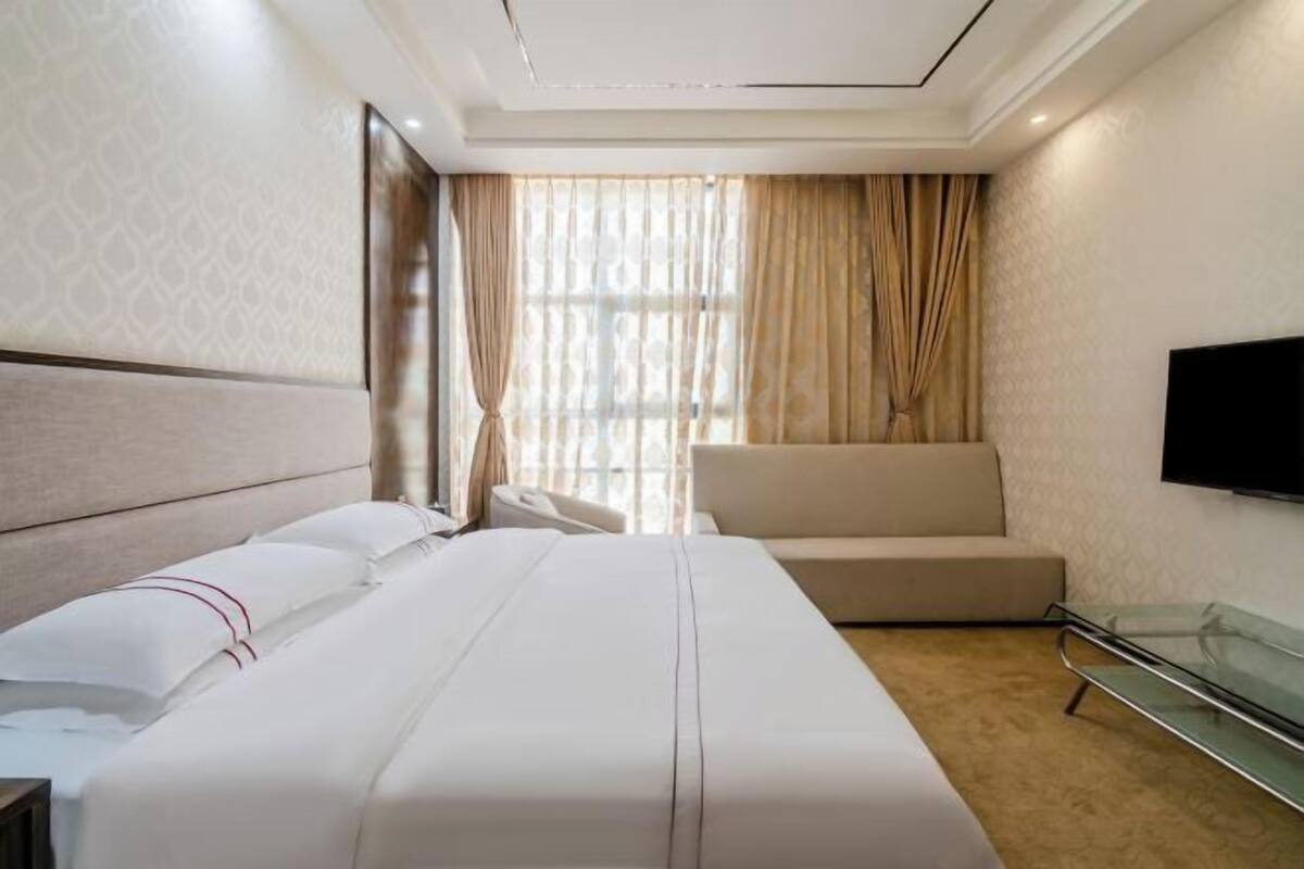 A hotel room with a large bed, a sofa, a TV mounted on the wall, and a window with curtains.
