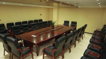 A conference room with a long table surrounded by chairs, a plant on the table, and a plain wall.