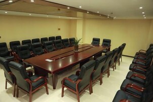 A conference room with a long table surrounded by chairs, a plant on the table, and a plain wall.