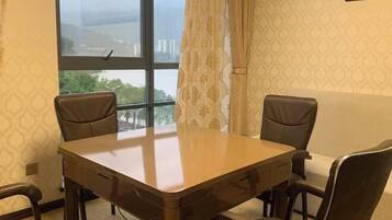 Quarto com mesa de madeira e quatro cadeiras, janela com vista para edifícios, papel de parede estampado e cortina.