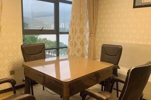 Quarto com mesa de madeira e quatro cadeiras, janela com vista para edifícios, papel de parede estampado e cortina.