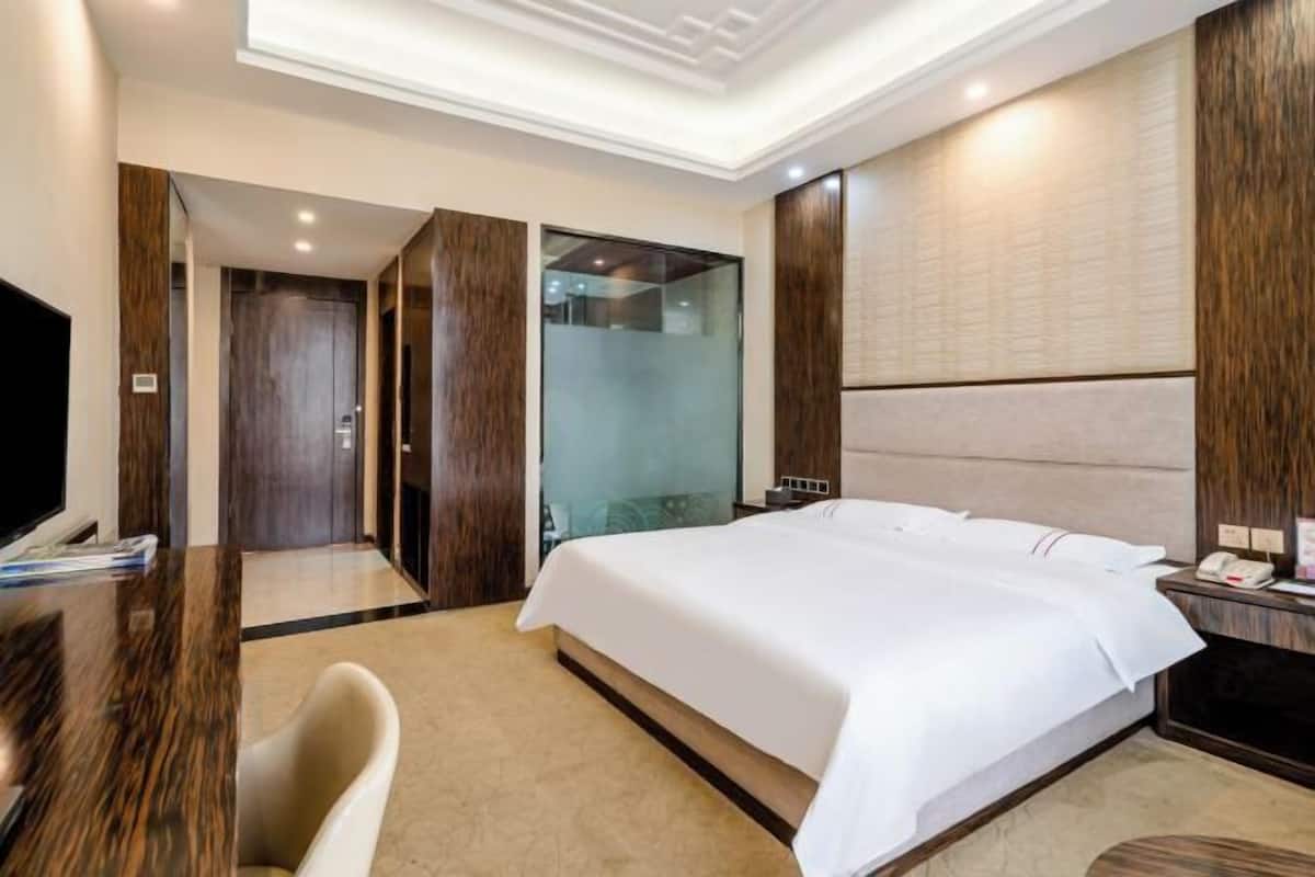A hotel room with a large bed, a desk with a chair, a TV, and a glass shower enclosure.