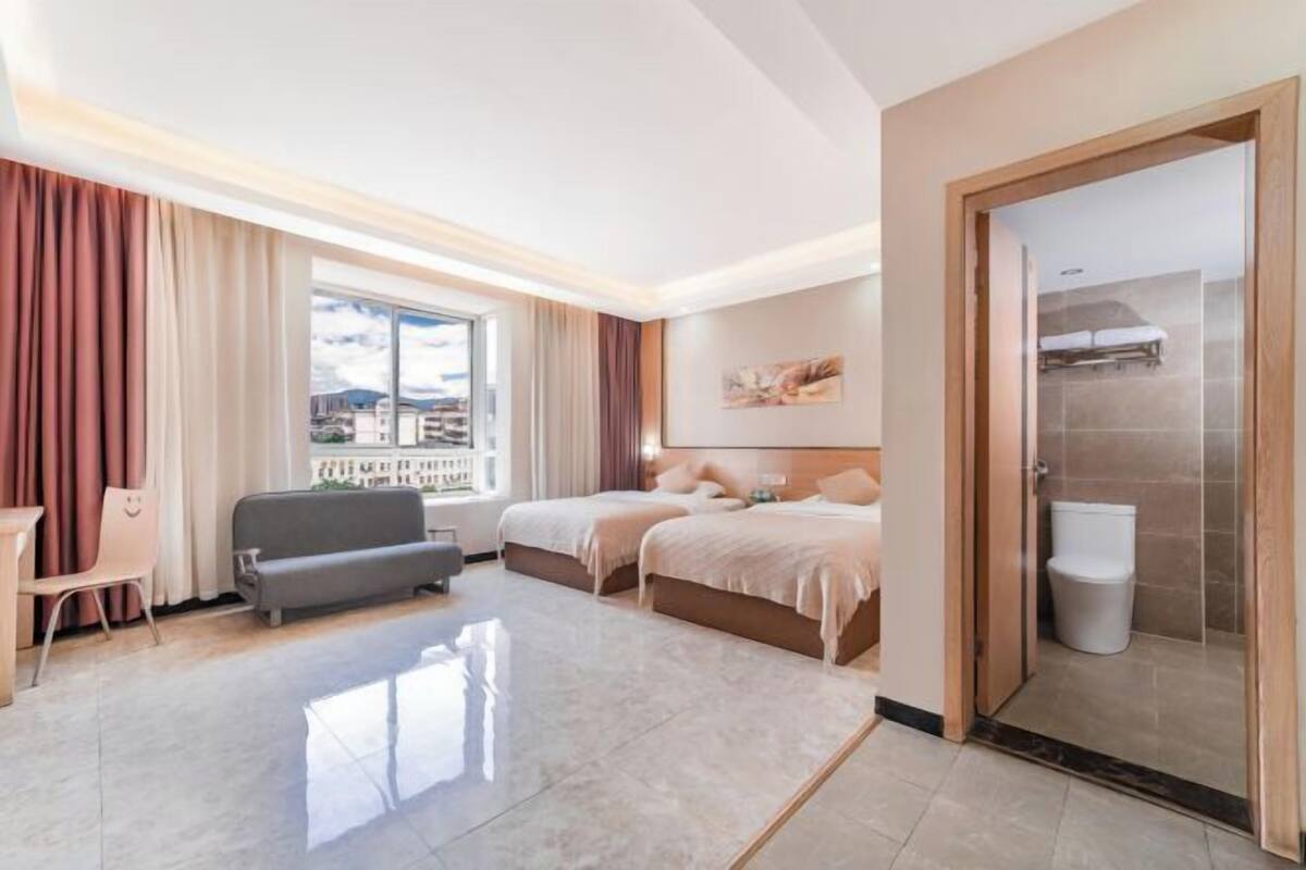 A hotel room with a large window, two beds, a sofa, a desk, and a bathroom.