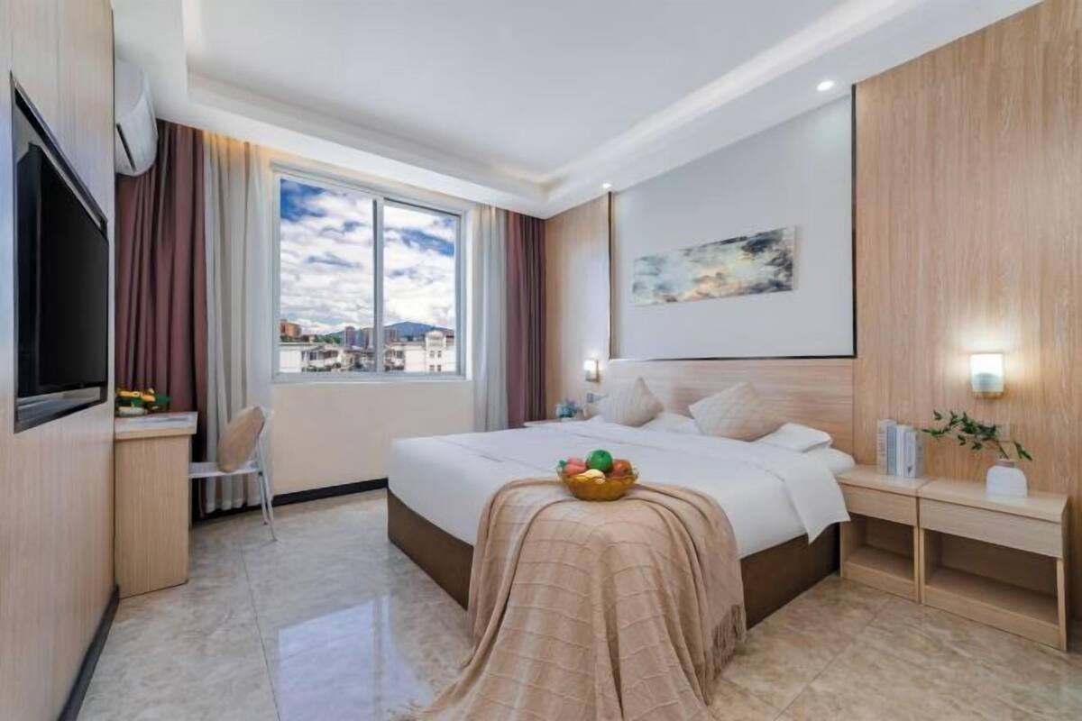 A hotel room with a bed, a desk, a chair, a TV, a window with a view, and a painting on the wall.