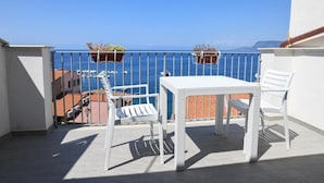 Un balcon avec une table et des chaises blanches, donnant sur une mer bleue et limpide et sur des bâtiments.