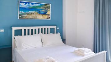 Une chambre avec un lit blanc, une tête de lit blanche et un tableau représentant des bateaux accroché au mur.