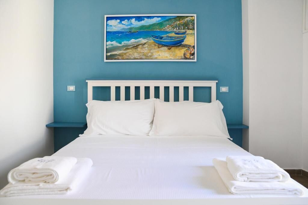 Una cama con ropa de cama y almohadas blancas, una pared azul y un cuadro de barcos en el mar.