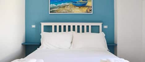 Una cama con ropa de cama y almohadas blancas, una pared azul y un cuadro de barcos en el mar.