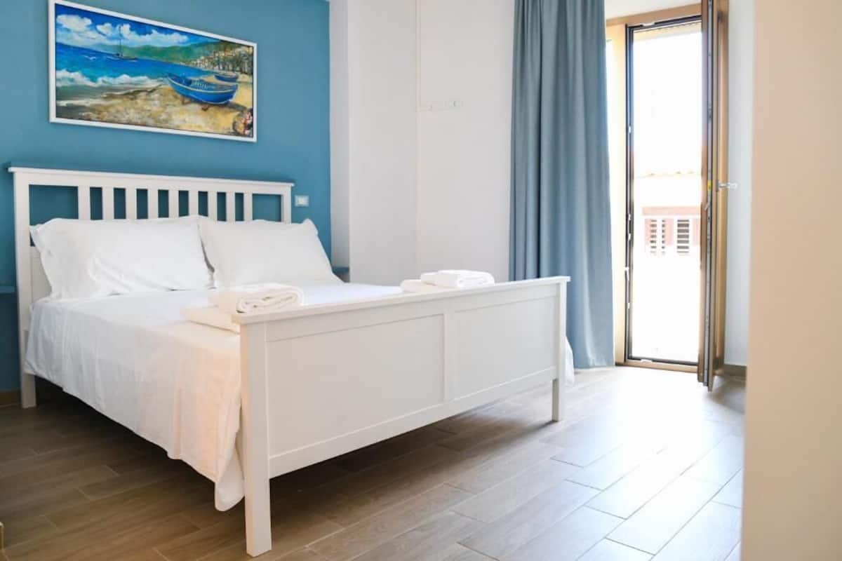 A bedroom with a white bed, a painting on the wall, and a view through a door.