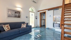 A modern living room with a blue sofa, a glass coffee table, and a kitchen area with a tiled backsplash. - Luxury sea View, With Infinity Pool in Sicily (Cefalù)