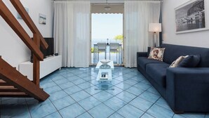 A living room with a blue tiled floor, a dark blue sofa, a white side table, a wooden staircase, and a balcony with a view. - Luxury sea View, With Infinity Pool in Sicily (Cefalù)