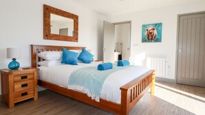 Una habitació amb un llit de fusta, roba de llit blanca, almoinades blaves, una làmpada blava i un quadre de una vaca.