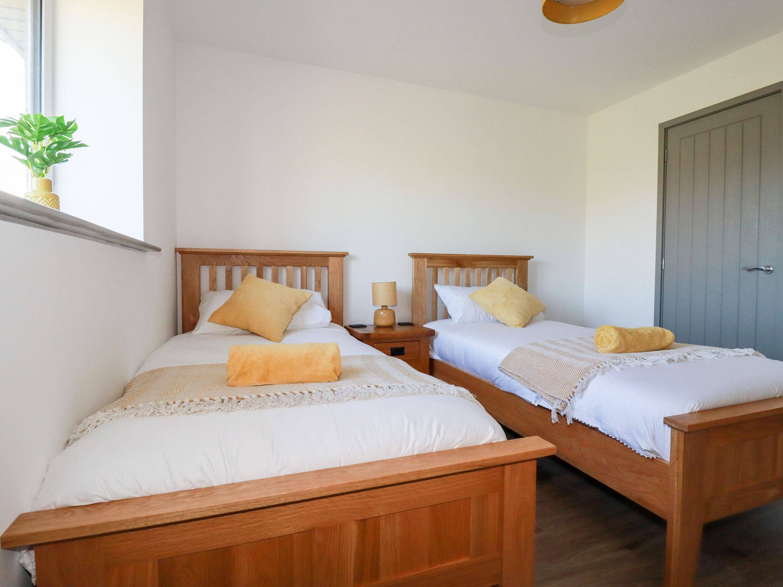 A room with two wooden beds, each with a white mattress and a yellow pillow. There is a small bedside table with a lamp on each bed.
