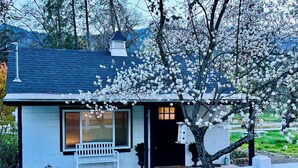 Une petite maison avec un porche, un banc et un arbre aux fleurs blanches.