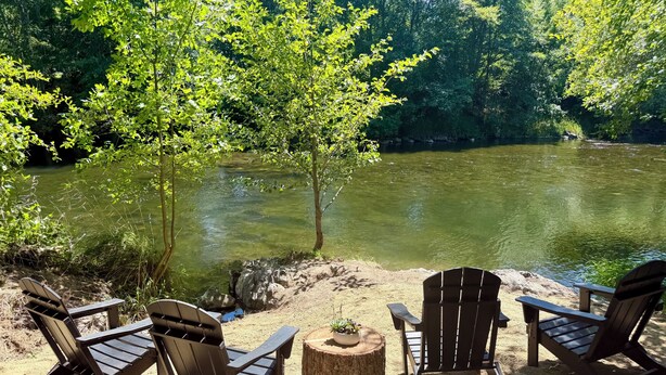 Un cadre paisible au bord de la rivière, avec trois fauteuils Adirondack, une petite table en bois et une vue dégagée sur la rivière, entourée d’une végétation luxuriante.