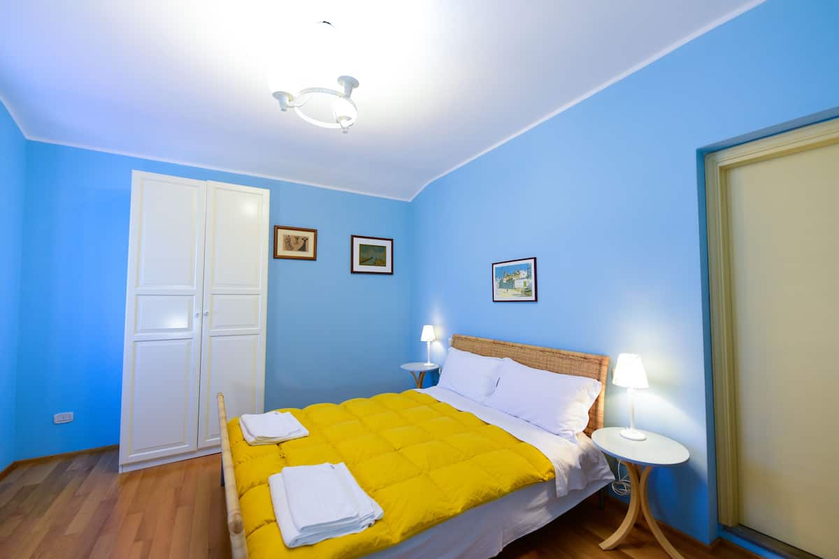 Een slaapkamer met een bed, witte gordijnen, een kledingkast en twee bedlampjes.