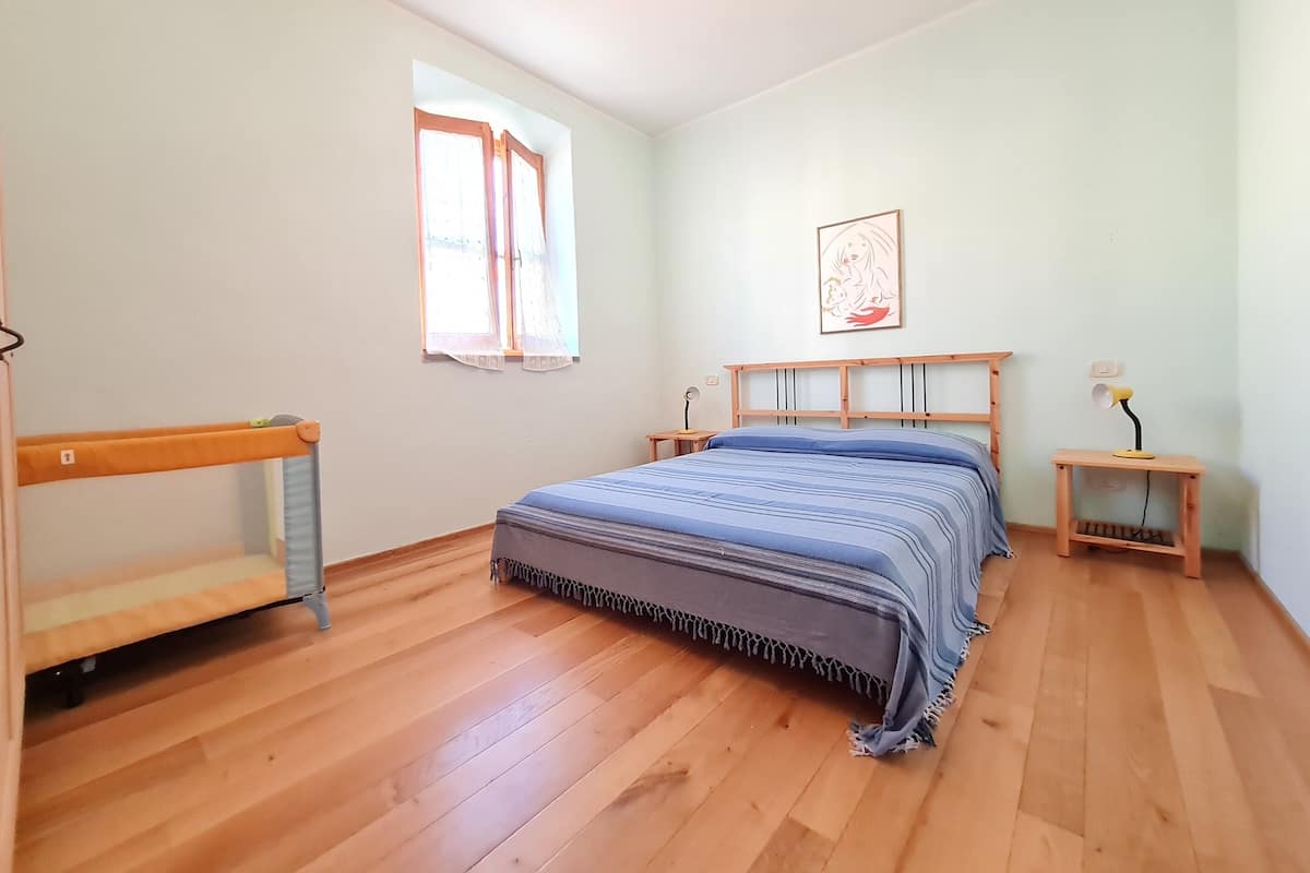 Een slaapkamer met een houten bed, een bankje, een raam met gordijnen, een nachtkastje met een lamp en een aan de muur hangend schilderij.