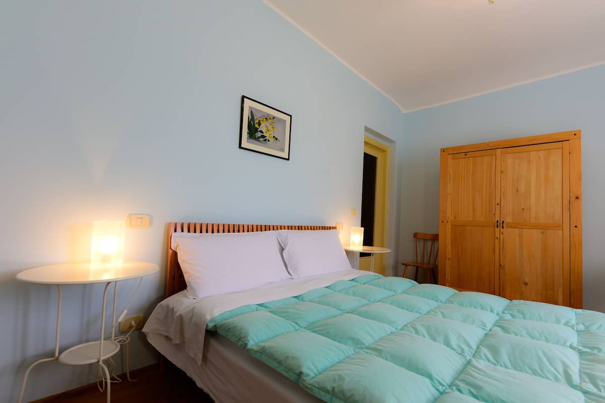 Een keurig opgemaakt bed met een lichtblauwe dekbedovertrek en witte kussens, een nachtkastje met een lamp, een houten garderobe en een ingelijst foto aan de muur.