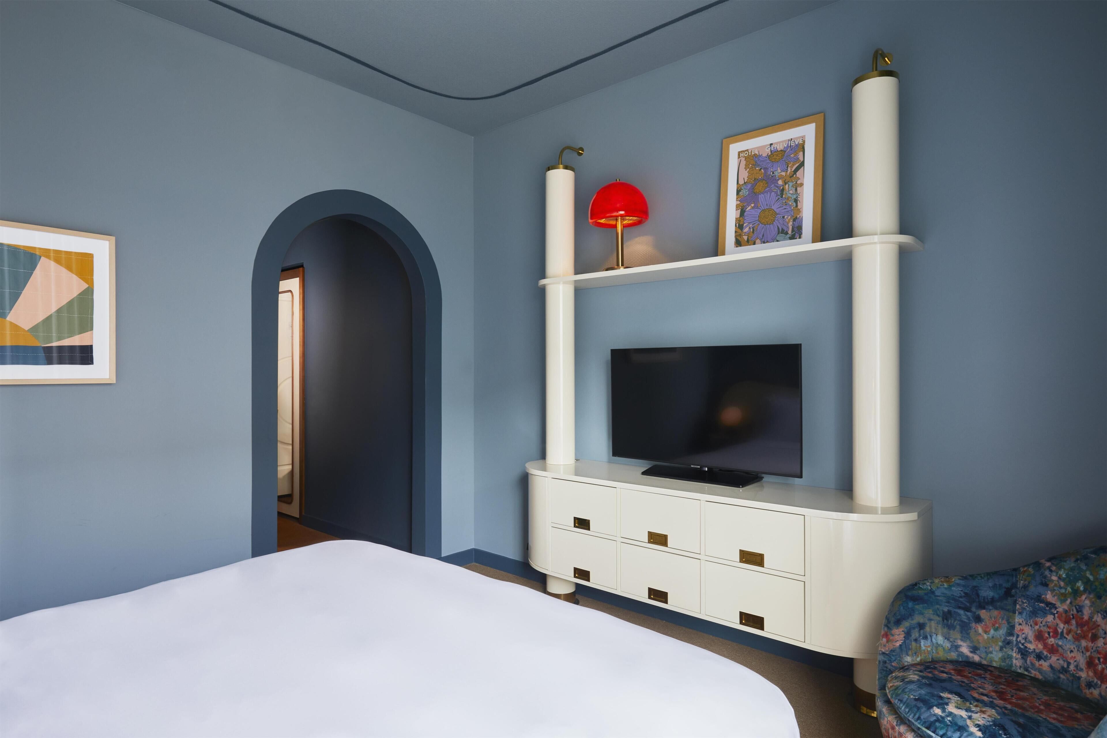 A hotel room with a bed, a TV on a white cabinet, a red lamp, and a floral chair.