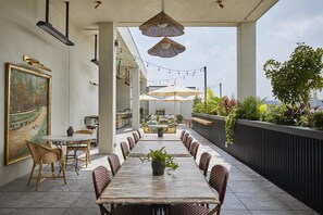 Un espace de restauration en plein air avec une longue table et des chaises en osier, un éclairage suspendu et une vue sur la ville.