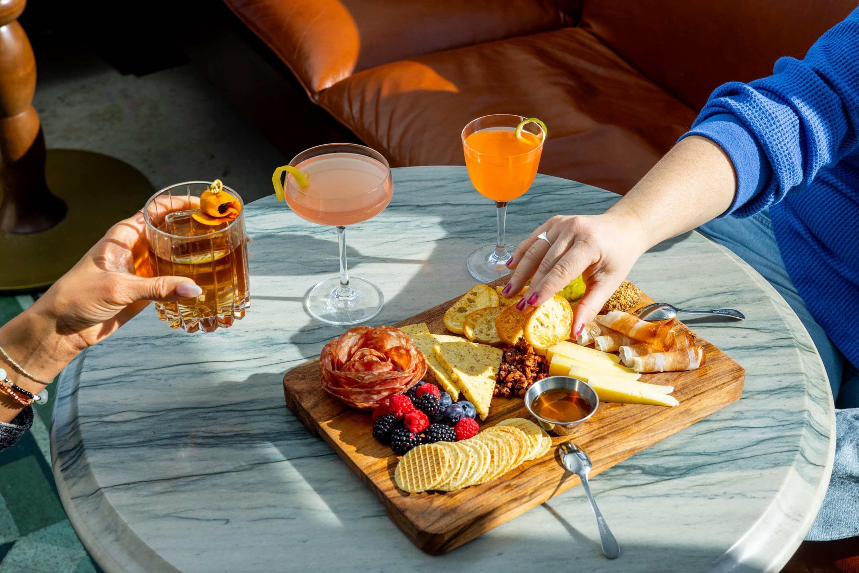 A marble table with a variety of food items and drinks, including cheese, fruits, and two cocktails.