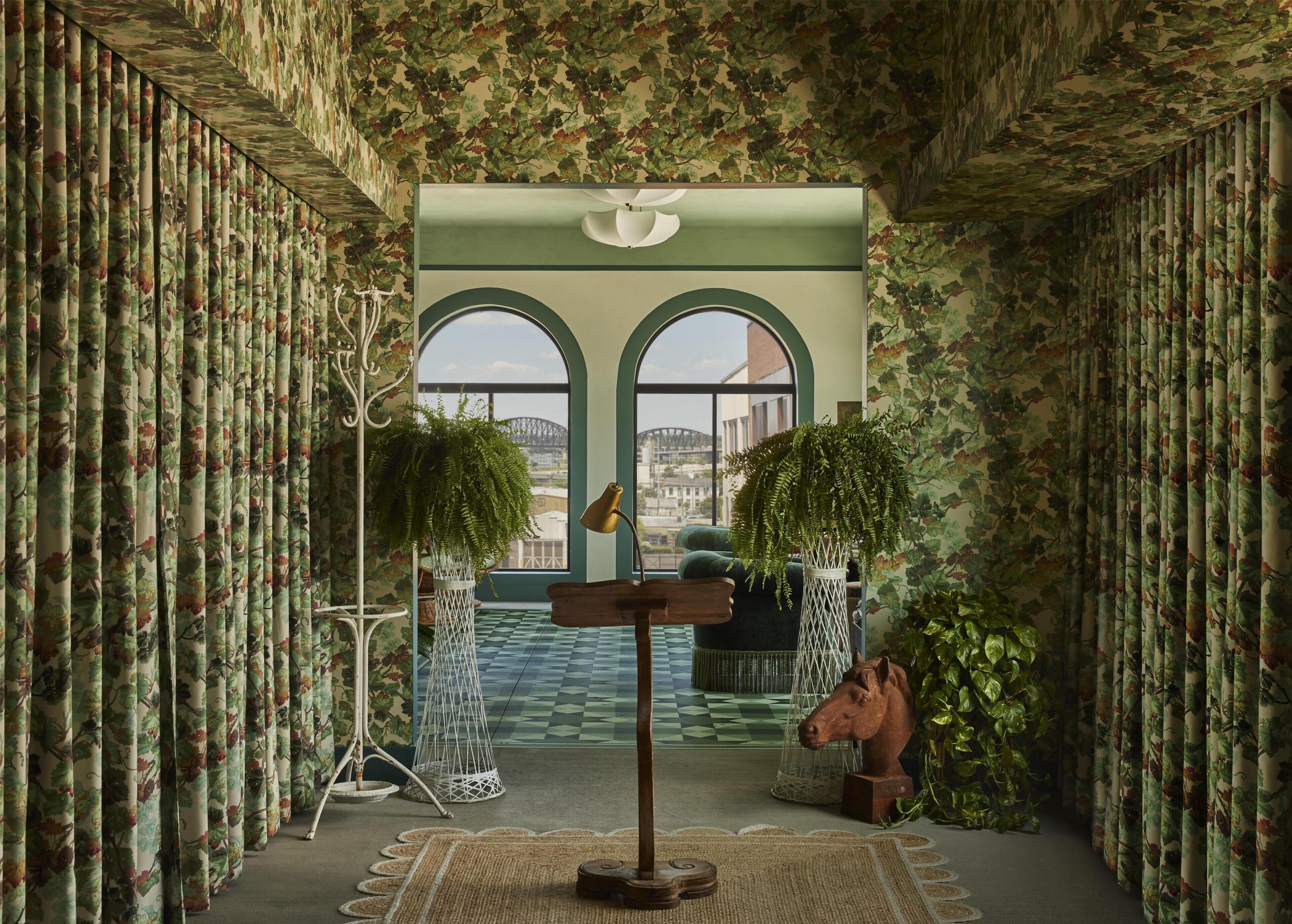 A room with patterned curtains, a large window with a view, a wooden sculpture, and a hanging plant.