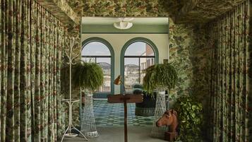 A room with patterned curtains, a large window with a view, a wooden sculpture, and a hanging plant.