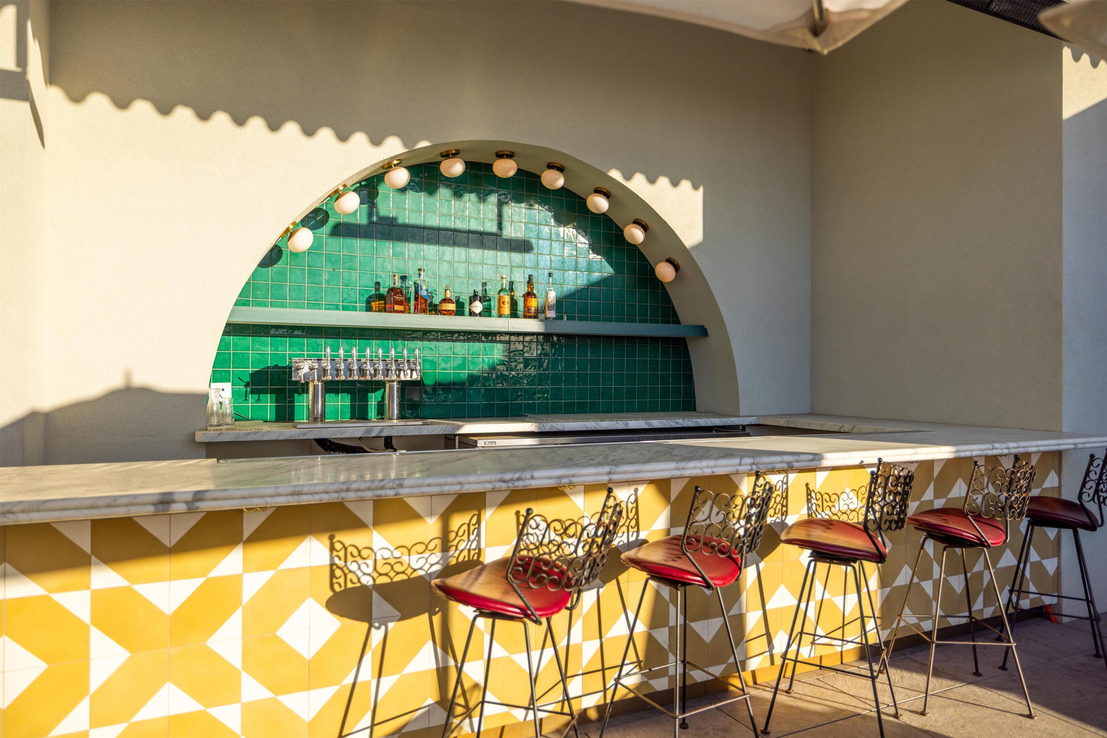 Un espace bar extérieur doté d’un mur recouvert de carreaux verts, d’un comptoir en marbre et de tabourets de bar en métal avec des coussins rouges.