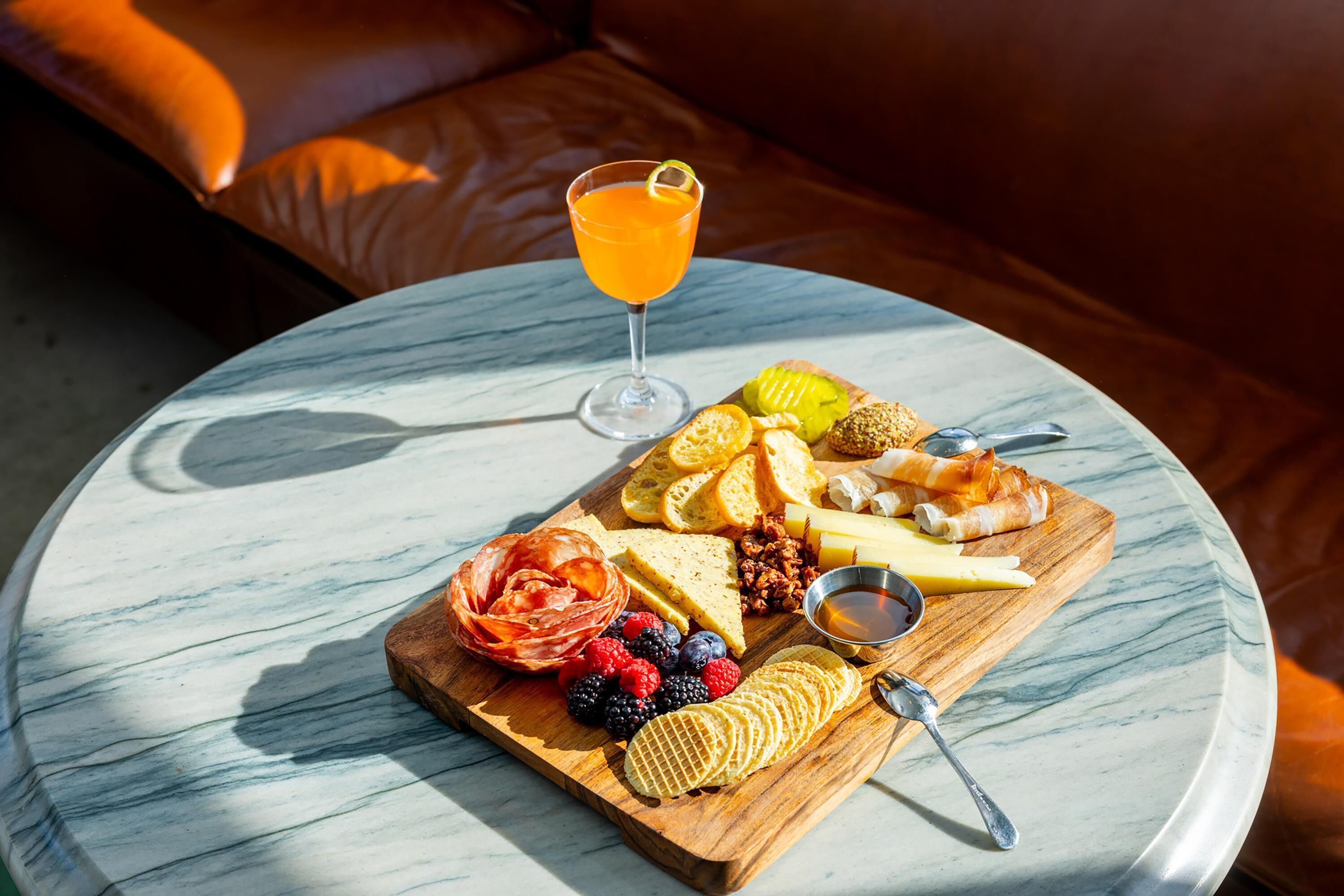 A marble table with a cheese board, fruits, nuts, and a glass of orange juice.