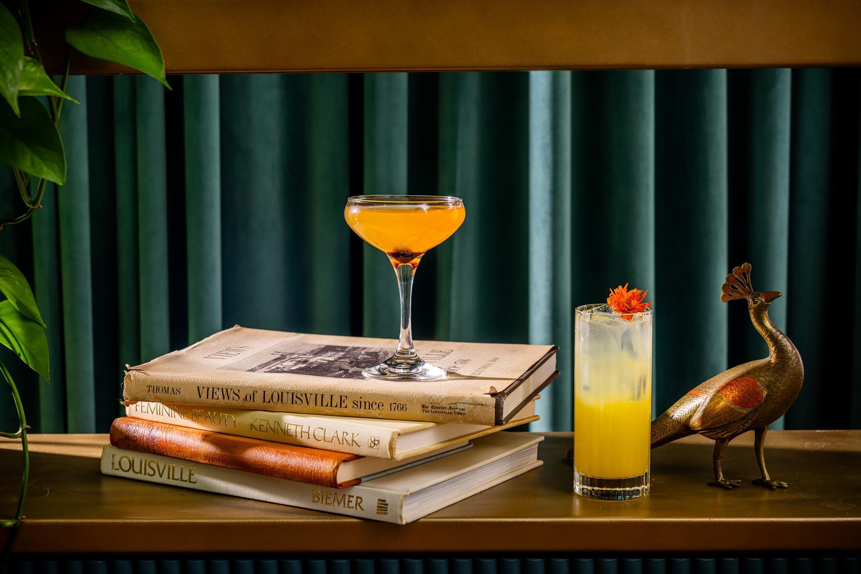 A stack of books, a martini glass with a cocktail, and a tall glass with a drink and a decorative item on a wooden surface against a green curtain backdrop.