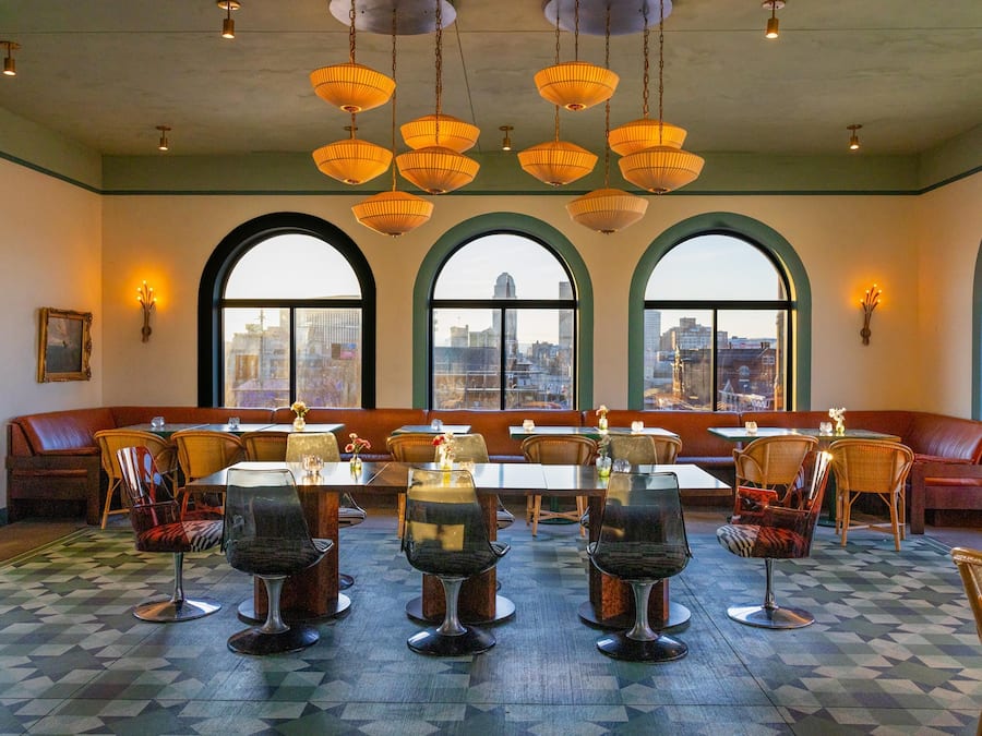 A restaurant with a long counter, multiple tables, and chairs. There are large windows with a view of the city.