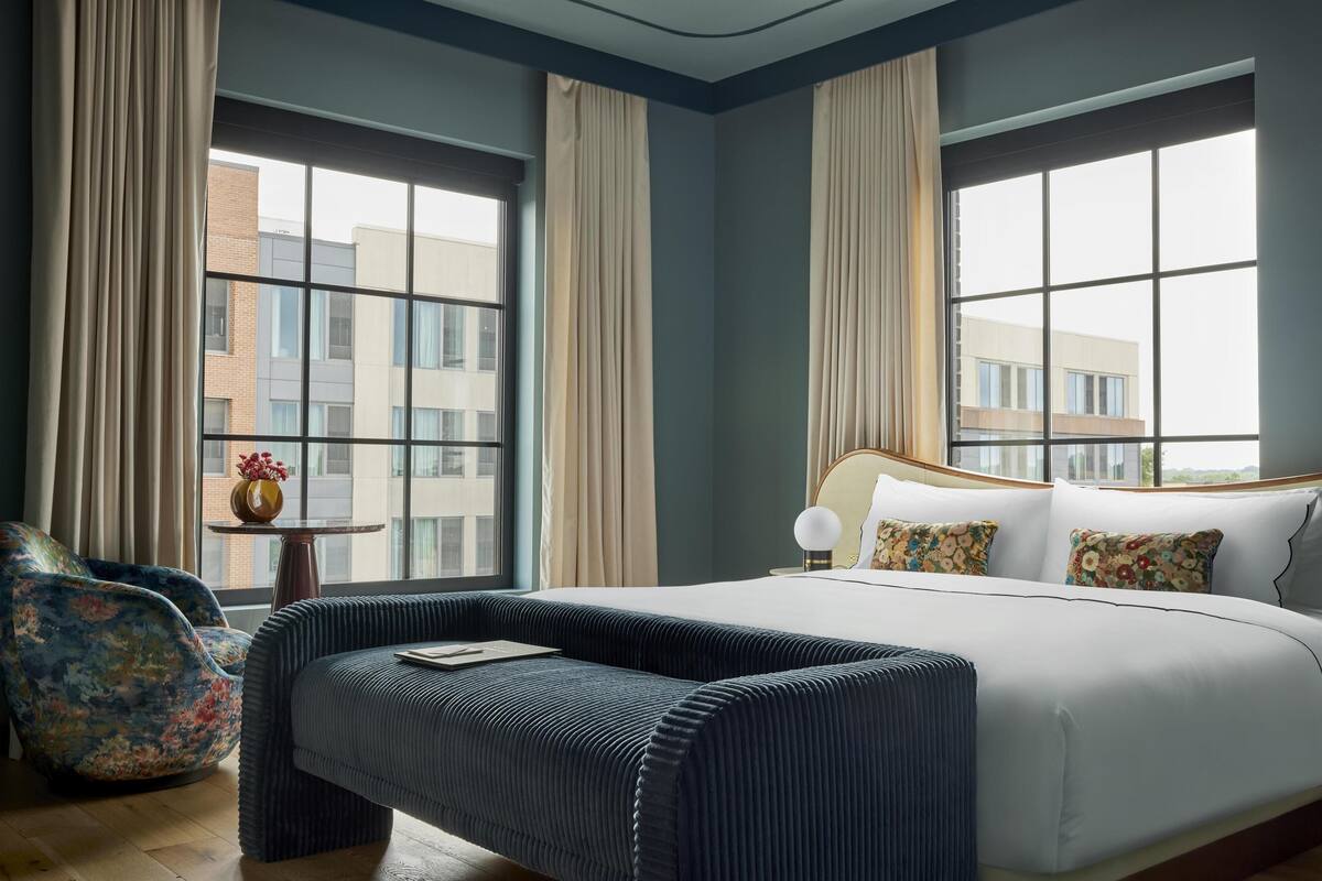 Ein modernes Hotelzimmer mit einem großen Bett, einer Sitzecke und Blick auf Gebäude durch große Fenster.
