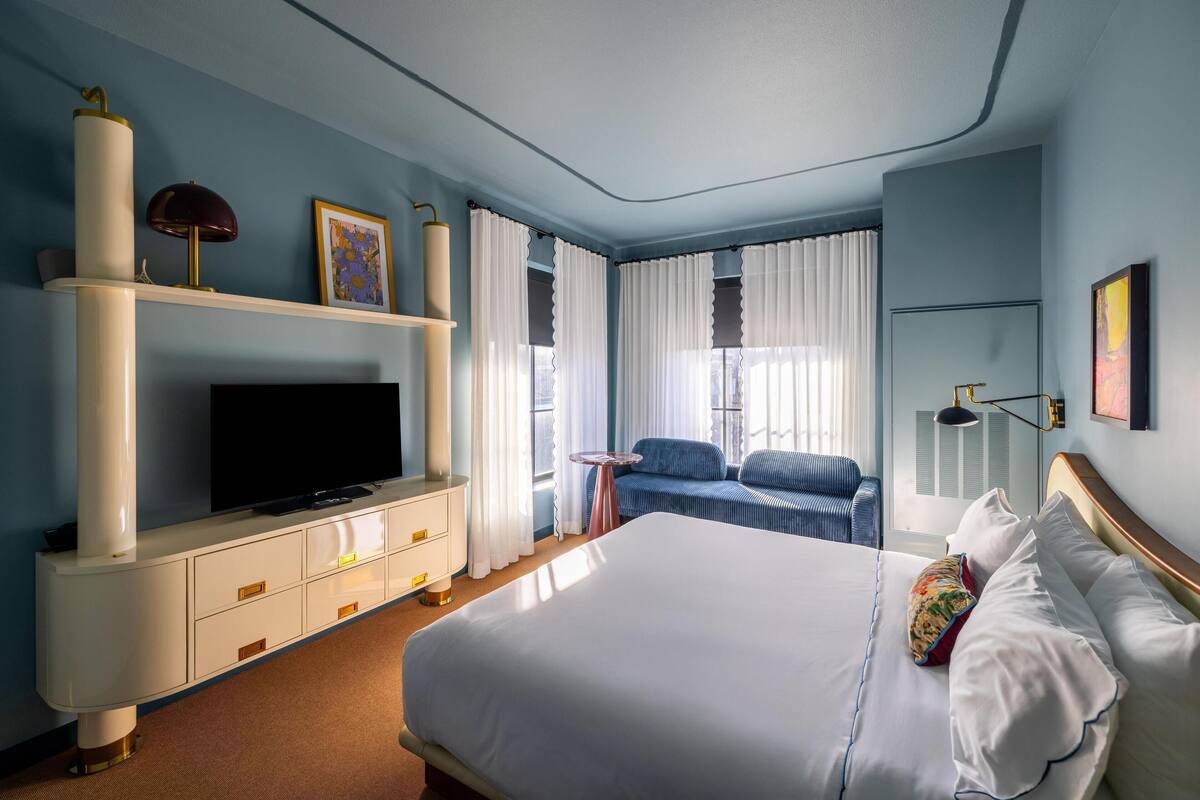 Ein Hotelzimmer mit einem großen Bett, einem Fernseher auf einer weißen Kommode, einem kleinen Tisch und einer Sitzecke mit Sessel und Sofa.