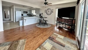 Uma sala de estar espaçosa com piso de madeira, uma ilha central com banquetas, um ventilador de teto, uma televisão grande e plana, e uma cozinha ao fundo.