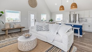 A modern living room with a white sofa, a round coffee table, and a kitchen area with a stainless steel refrigerator. - Reel Paradise: NEW Luxury 5BR! Private POOL + FirePit & OutdoorTV. Great Views! (Port Aransas)