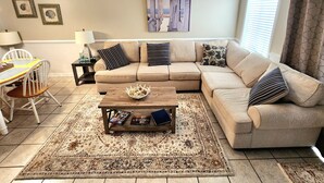 Ein Wohnzimmer mit einem beigefarbenen Ecksofa, einem Holztisch, einem gemusterten Teppich und einem Deckenventilator.