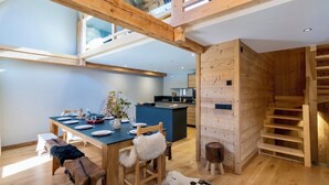 Una cocina moderna con vigas de madera, un comedor con una mesa dispuesta para cuatro personas, y una escalera que conduce a un nivel superior.