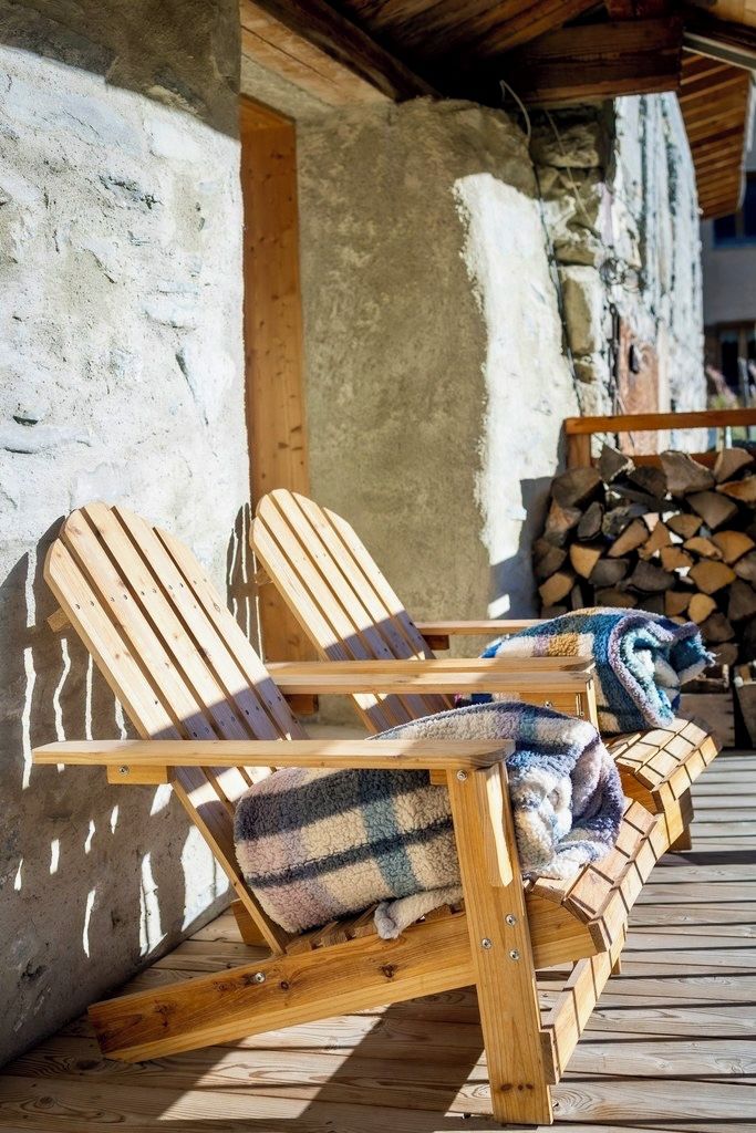 Two wooden lounge chairs with blankets, a stack of firewood, and a rustic stone wall.