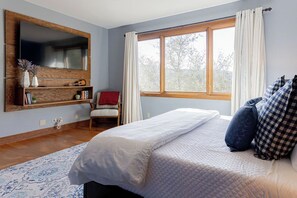 A bedroom with a large bed, a TV mounted on the wall, a window with curtains, and a chair.