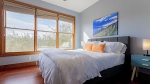 Ein Schlafzimmer mit einem großen Fenster, einem Bett mit weißer Bettwäsche, einem blauen Nachttisch und einem Landschaftsbild an der Wand.