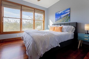Une chambre avec une grande fenêtre, un lit recouvert de draps blancs, une table de chevet bleue et un tableau représentant un paysage accroché au mur.