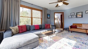 Ein Wohnzimmer mit einem Ecksofa, einem Couchtisch, einem gemusterten Teppich, einem Deckenventilator und einer Treppe, die eine weitere Ebene hinaufführt.