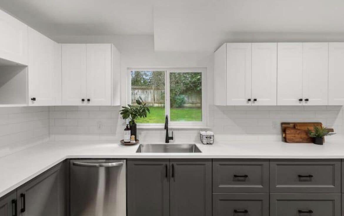 A modern kitchen with white cabinets, a stainless steel sink, and a view of a backyard through a large window.
