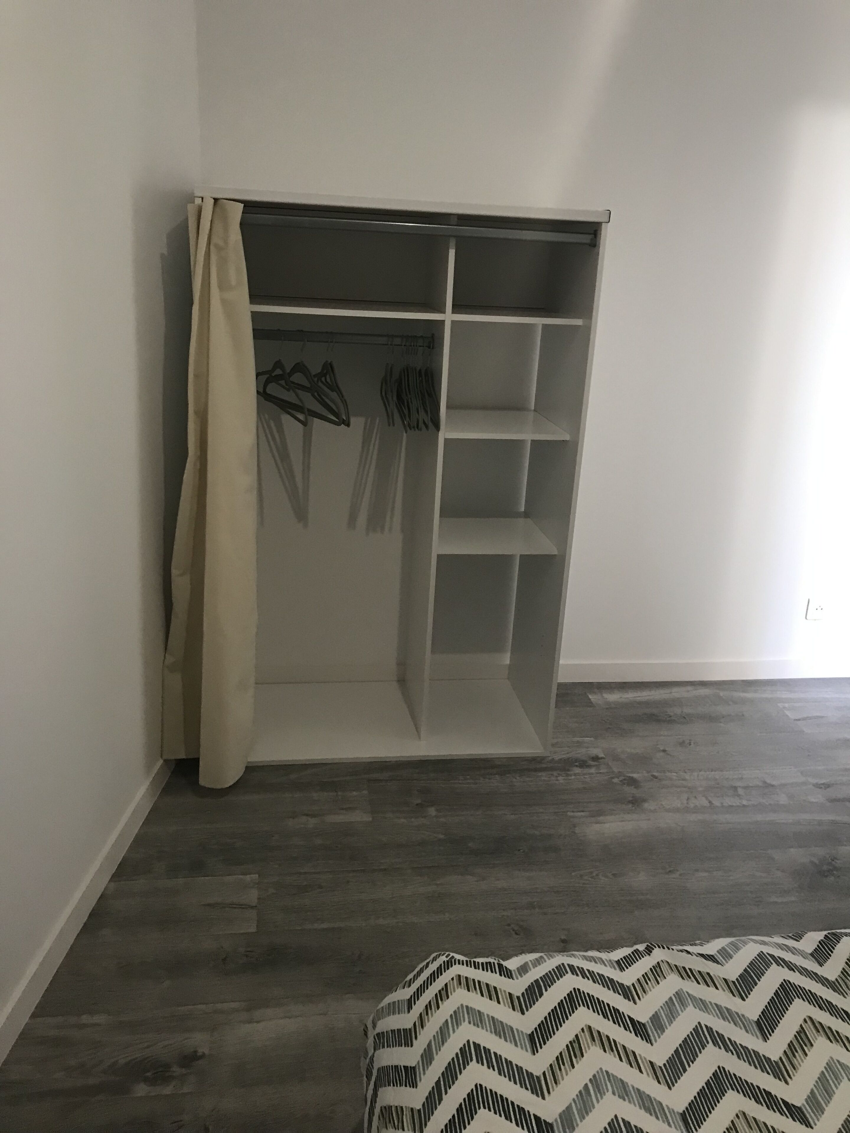 Een inloopkast met hangruimte en planken, een bed met een bedovertrek met dessin, en een houten vloer.
