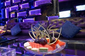 Eine Bar mit einem Glastisch, der mit Früchten dekoriert ist, und einem violetten, gepolsterten Sofa mit blauen Kissen.